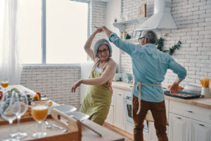 Playful senior couple in aprons dancing and smiling while preparing dinner at home.