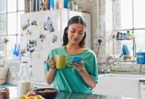 Woman using a financial app on her cellphone while enjoying a cup of coffee in her kitchen at home.