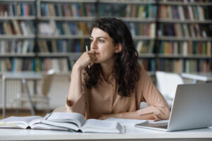Young woman looks pensive and thoughtful in front of computer and books.