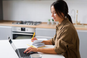Young woman writes notes on paper while researching on her laptop.