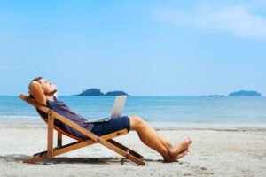 Smiling man with computer on his lap, leaning back in a lounge chair and relaxing on the beach.