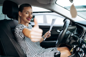 Smiling woman sitting in a car and holding a contract with one hand, her other hand giving the thumb's up signal.