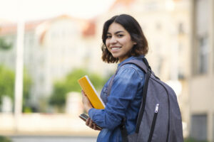 Happy Middle Eastern young woman posing outside at university campus, holding notebook.