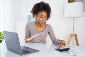 Black woman managing budget with laptop and calculator at home table.