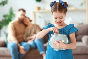 Little girl in pigtails adding money to a piggy bank while her parents watch in the background.