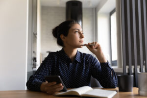Pensive young Indian woman with notebook and cellphone, looking off into the distance.