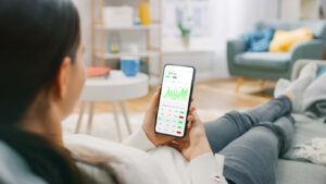 Young woman at home sitting on a couch and using smartphone with stock market app.