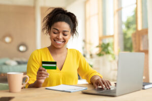 Young Black woman holding credit card and using her computer at a desk at home.