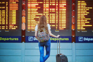 Young tourist girl with backpack and carry-on luggage in international airport, looking at flight information board with her back to camera.