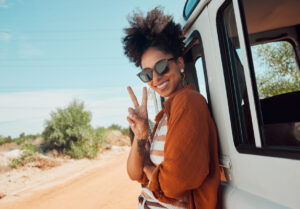 Relaxed Black woman in sunglasses on vacation, leaning against a van and giving a peace sign.