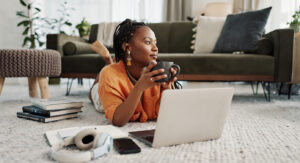 Young Black woman lounging on the floor in living room of modern apartment, with a coffee and her laptop.
