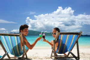 Couple sitting in loungers on a beach, cheersing each other with cocktails.