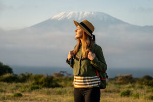 A young woman In a hat stands against the backdrop of the Kilimanjaro volcano and looks away.