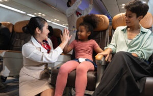 Flight attendant high-fives young passenger while child's mother watches and smiles.
