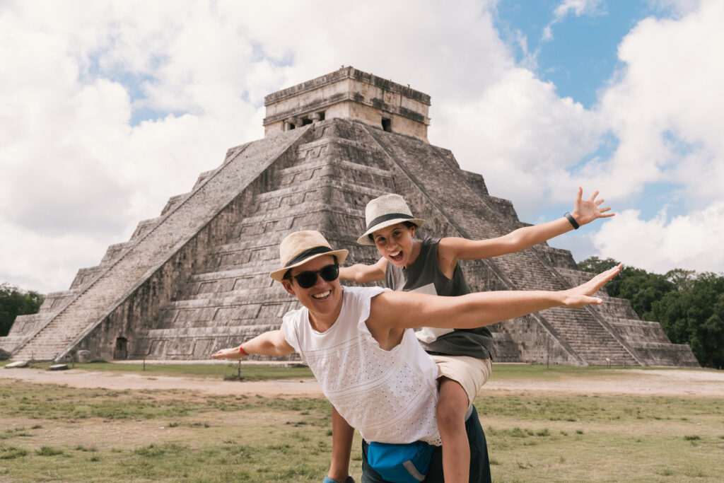 Parent with child on their back, standing with arms spread in front of Chichen Itza ruins in Mexico