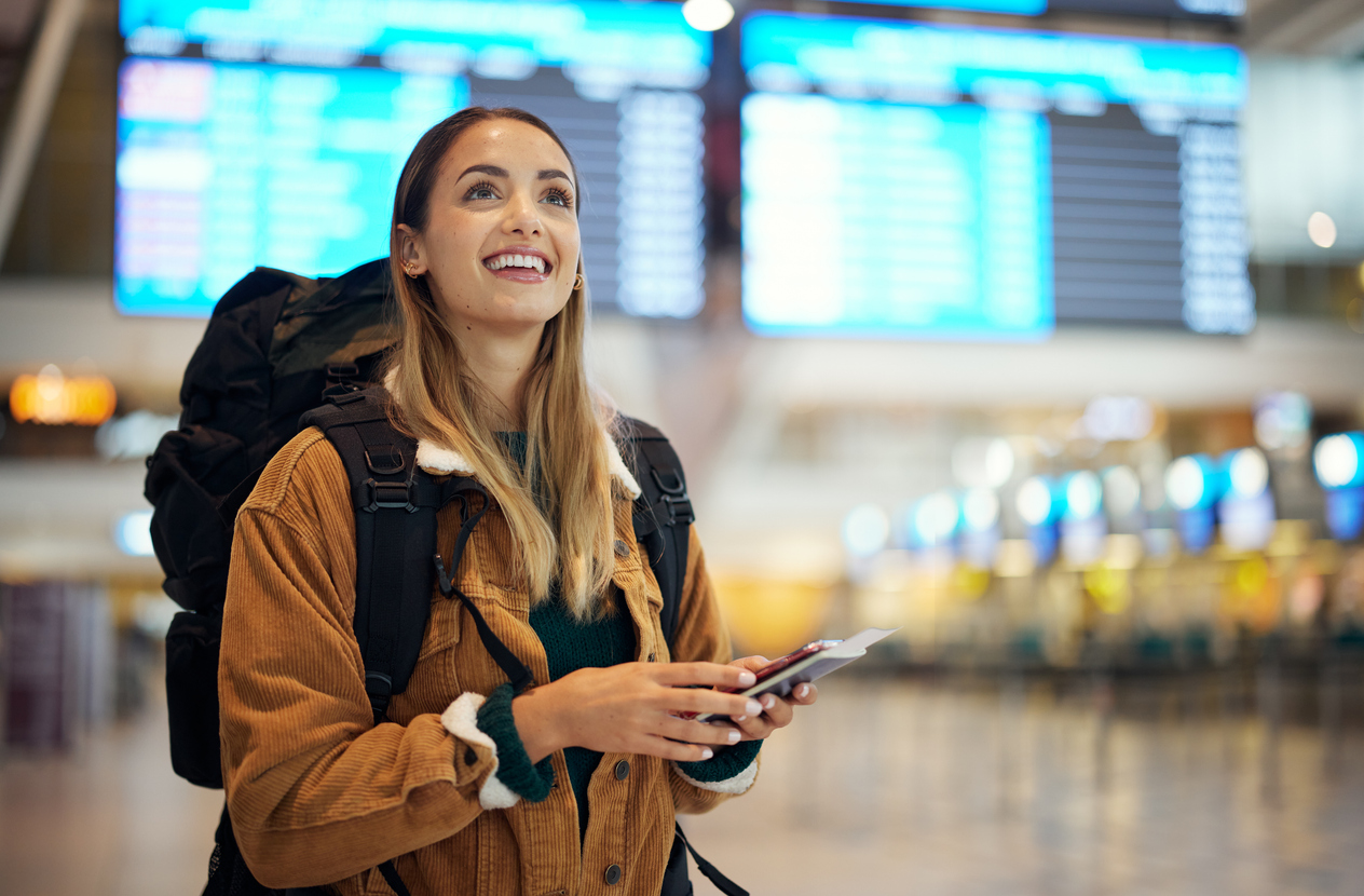 Young woman standing in the airport near the departures board, wearing a backpack and holding her passport and ticket