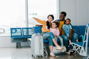 Smiling family with baggage and kids sitting in airport while mother pointing at something in distance.