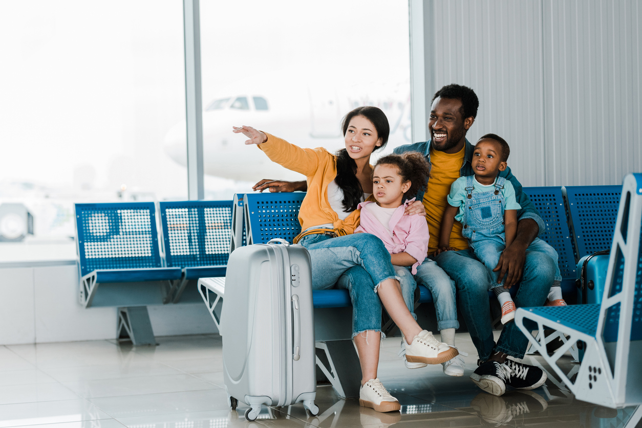 Smiling family with baggage and kids sitting in airport while mother pointing at something in distance.