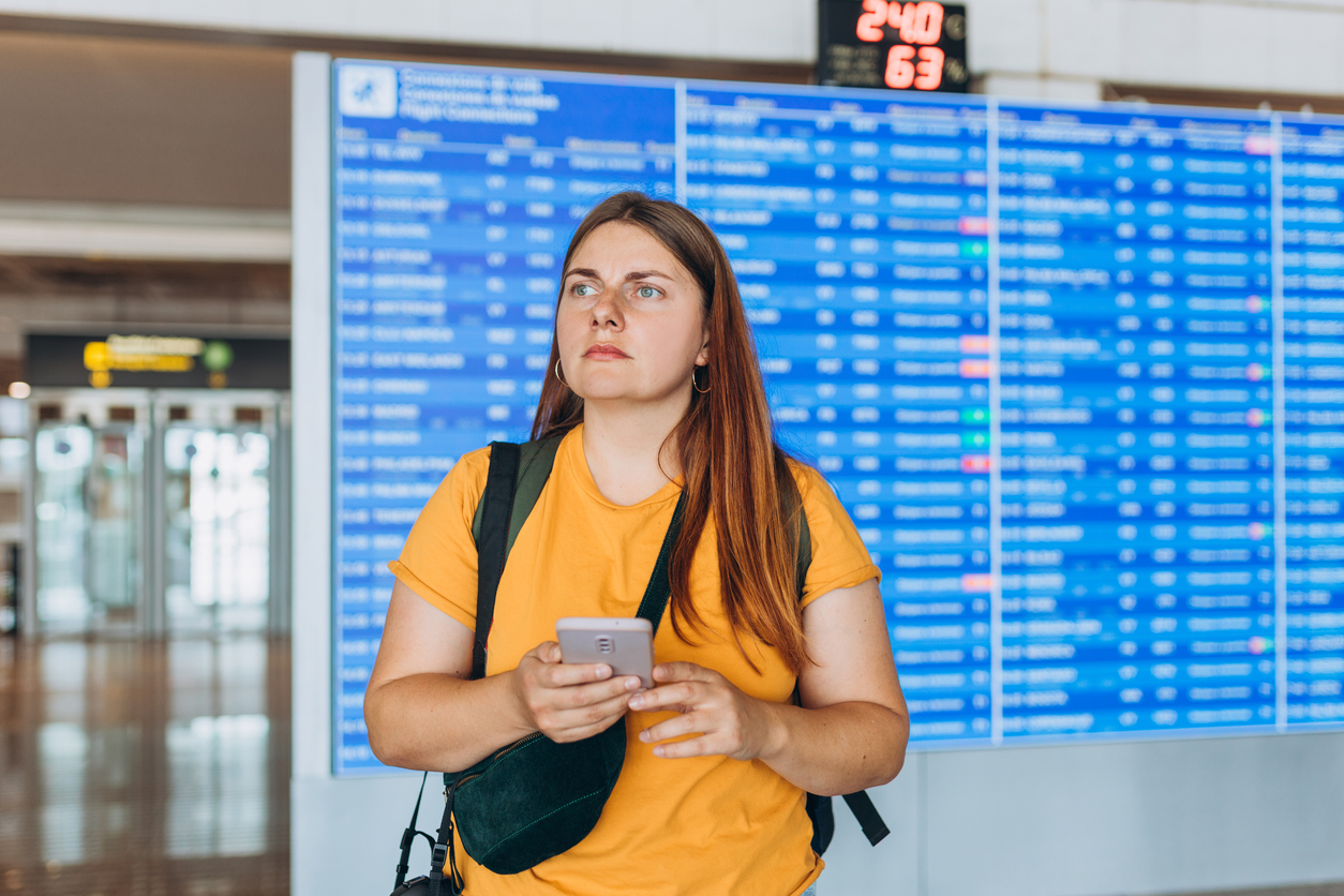 Perplexed-looking woman holding cellphone in front of the departures board at an airport