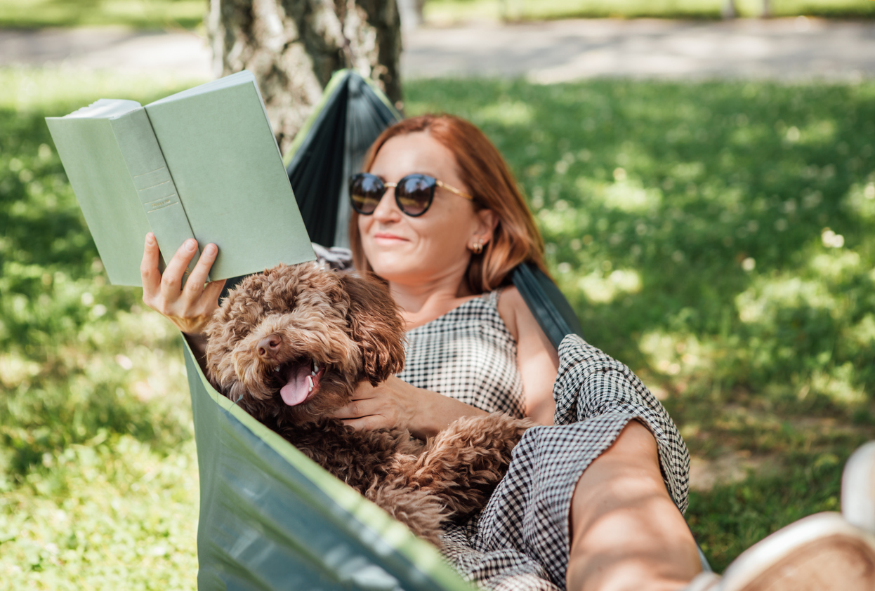 Woman relaxing in a hammock, reading a book and holding her dog