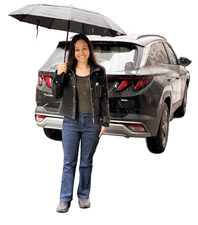 Smiling woman stands in front of a gray SUV, holding a black umbrella in the rain.